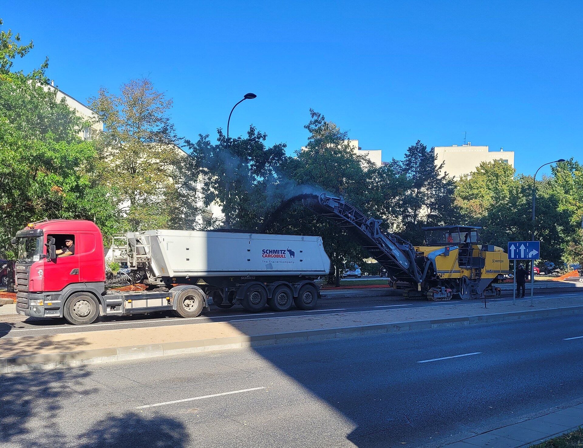Three-axle Schmitz Cargobull tipper semi-trailer behind a red Scania tractor, being loaded with millings by a cold milling machine during road resurfacing in Warsaw